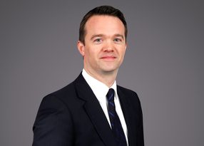 Head-and-shoulders portrait of an individual in a dark suit, white shirt and blue-striped tie, smiling slightly against a grey gradient background.
