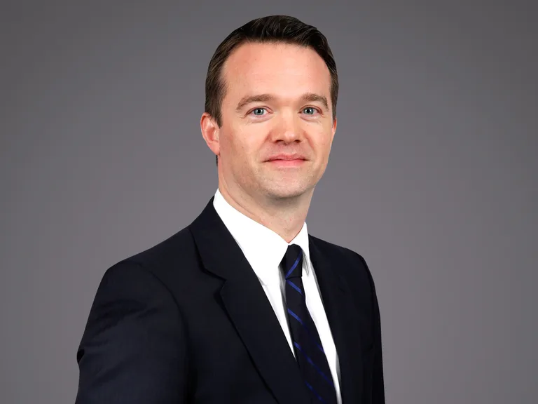 Head-and-shoulders portrait of an individual in a dark suit, white shirt and blue-striped tie, smiling slightly against a grey gradient background.