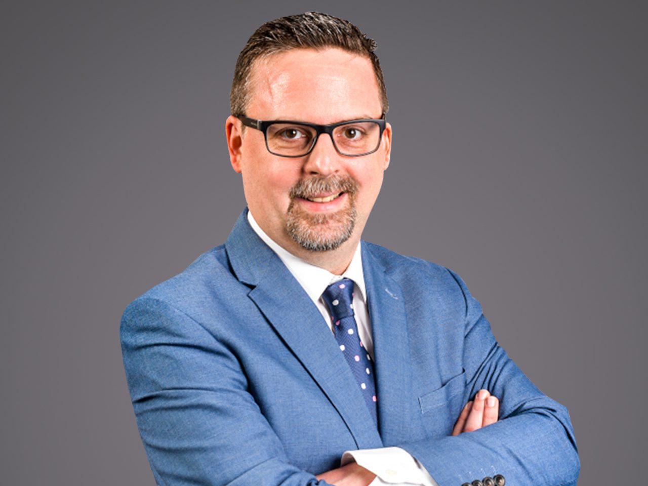 Portrait of a person in a blue suit, white shirt and polka-dot tie, wearing glasses with short hair and a trimmed beard, arms crossed against a grey gradient backdrop.