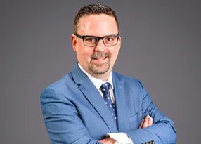 Portrait of a person in a blue suit, white shirt and polka-dot tie, wearing glasses with short hair and a trimmed beard, arms crossed against a grey gradient backdrop.
