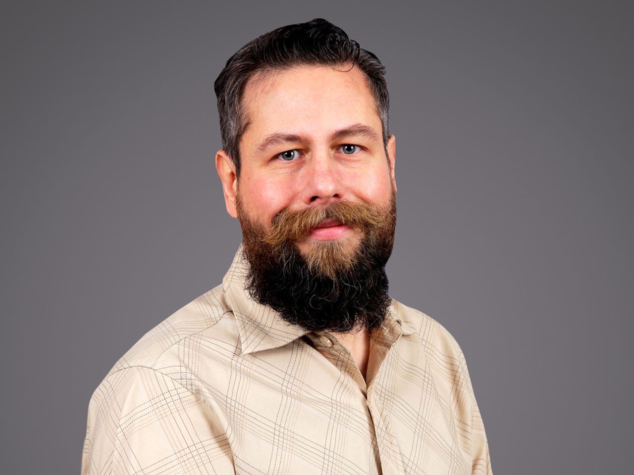 Portrait of a person with dark hair and a full beard, wearing a beige plaid shirt, posed against a plain grey studio background.