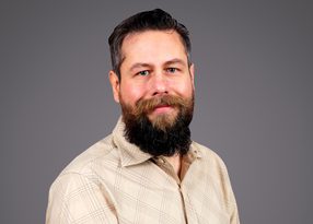 Portrait of a person with dark hair and a full beard, wearing a beige plaid shirt, posed against a plain grey studio background.