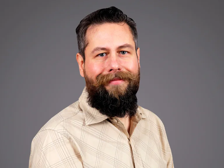Portrait of a person with dark hair and a full beard, wearing a beige plaid shirt, posed against a plain grey studio background.