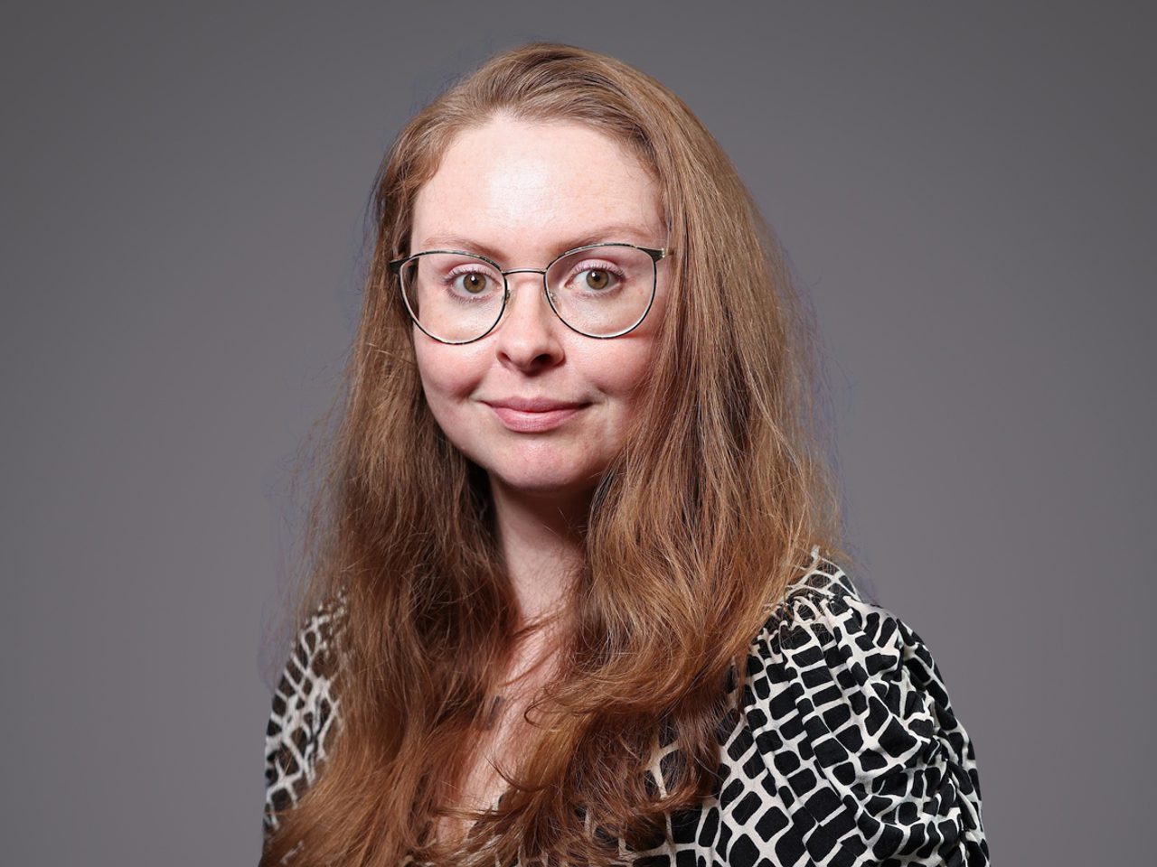 Portrait of a person with long light brown hair, round glasses, and a black-and-white patterned top, smiling softly against a grey background.