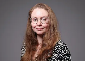 Portrait of a person with long light brown hair, round glasses, and a black-and-white patterned top, smiling softly against a grey background.