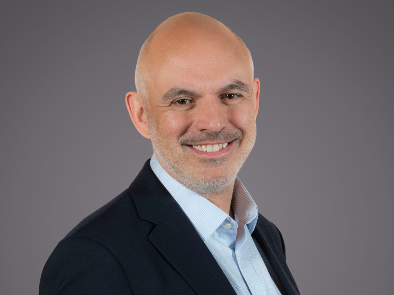 Bald person in a dark blazer and light blue shirt, smiling, against a plain grey studio background.