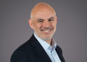 Bald person in a dark blazer and light blue shirt, smiling, against a plain grey studio background.