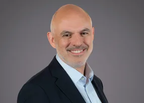 Bald person in a dark blazer and light blue shirt, smiling, against a plain grey studio background.