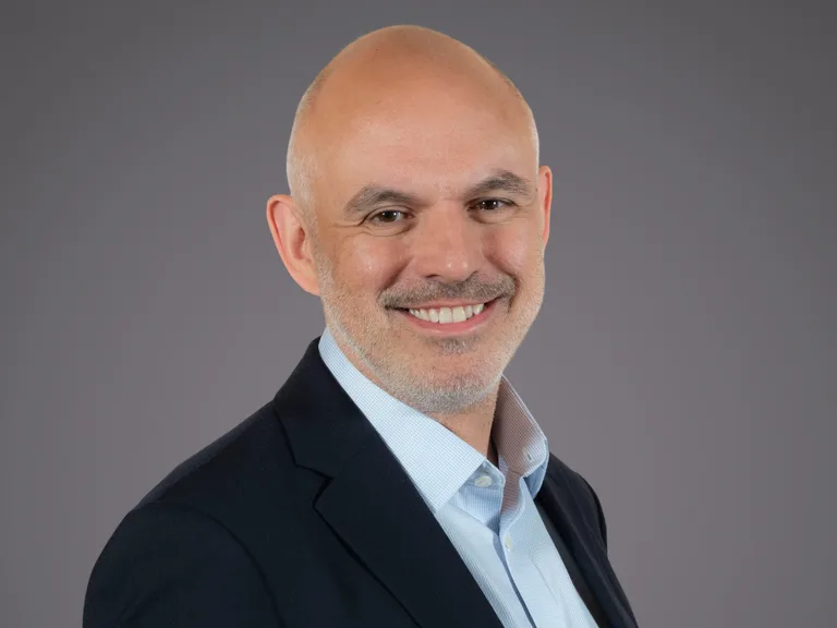 Bald person in a dark blazer and light blue shirt, smiling, against a plain grey studio background.