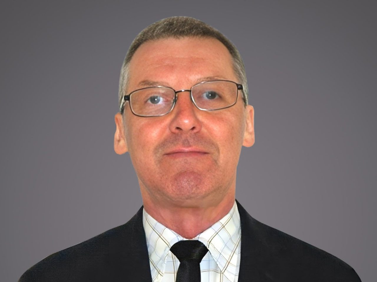 Persona adulta con capelli corti grigi e occhiali, indossa giacca scura, camicia a quadri e cravatta nera; sfondo grigio uniforme, espressione neutra.