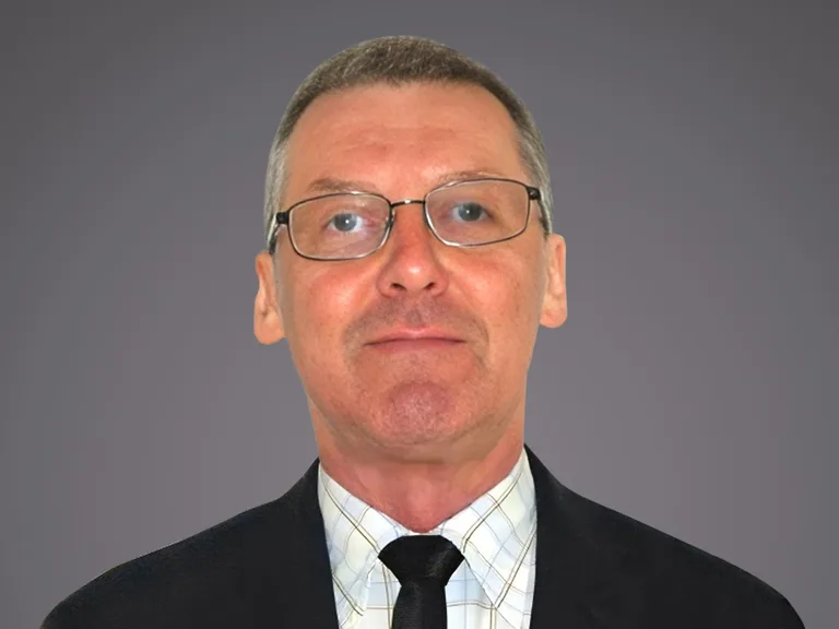 Individu d'âge moyen, cheveux gris courts, portant des lunettes rectangulaires, chemise à carreaux claire et cravate noire, sur fond gris uniforme.