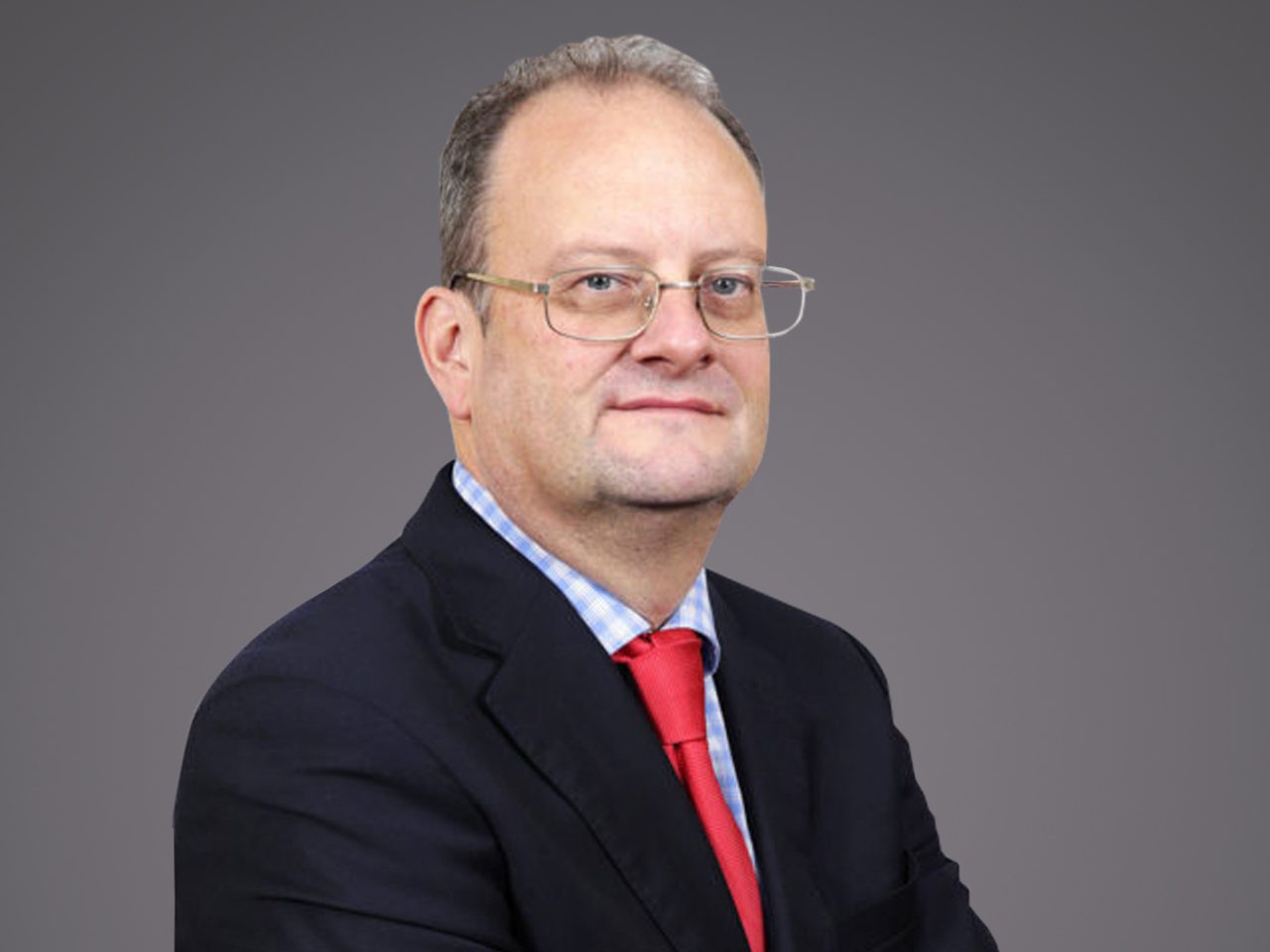 Portrait of a person in a dark suit, light blue checked shirt and red tie, wearing glasses, posed against a grey backdrop.
