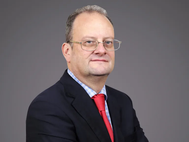 Portrait of a person in a dark suit, light blue checked shirt and red tie, wearing glasses, posed against a grey backdrop.