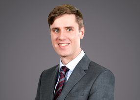 Portrait of a person in a grey suit and striped burgundy tie, smiling at the camera against a plain grey background.