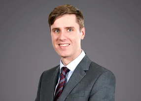 Portrait of a person in a grey suit and striped burgundy tie, smiling at the camera against a plain grey background.