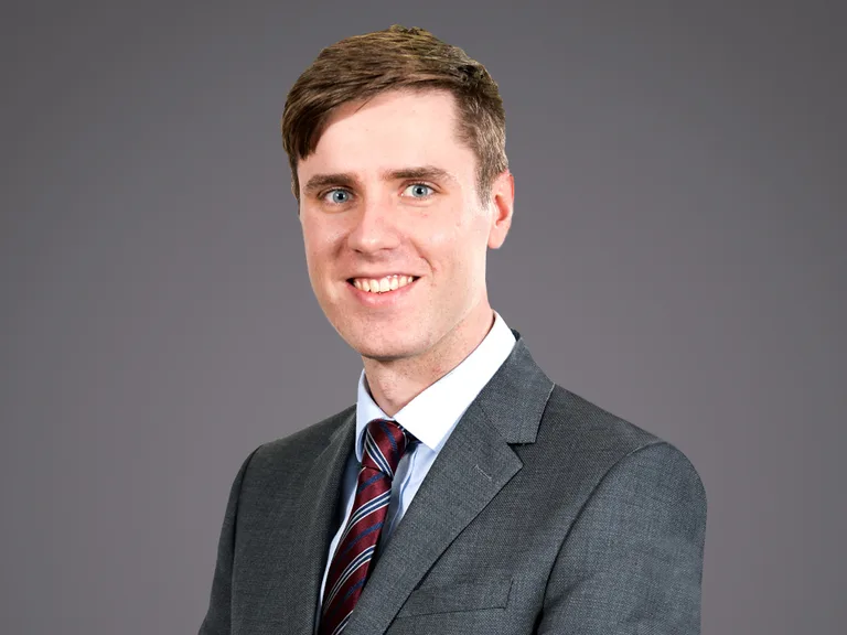 Portrait of a person in a grey suit and striped burgundy tie, smiling at the camera against a plain grey background.