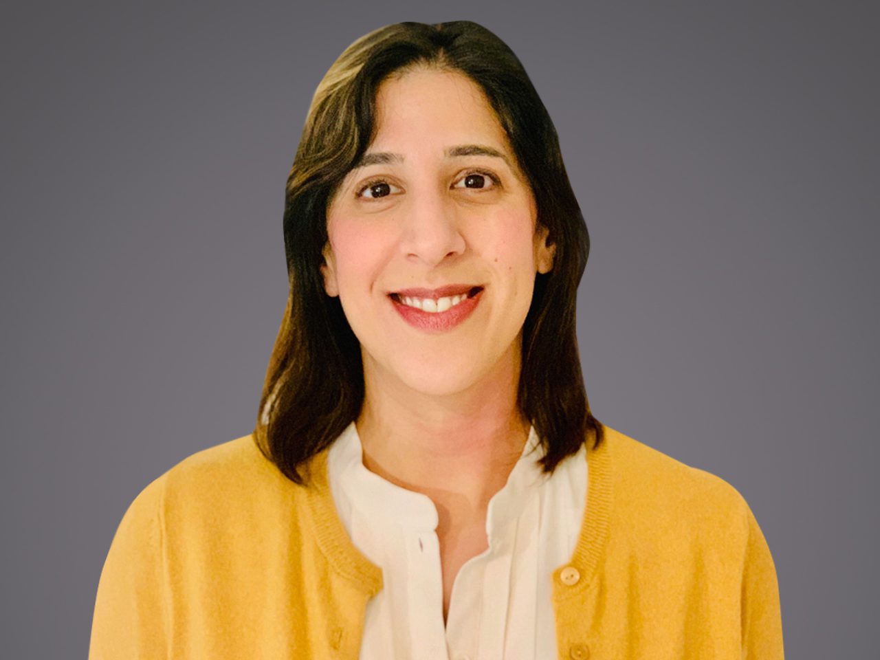 Portrait of a person with shoulder-length dark hair, smiling, wearing a white blouse and yellow cardigan, against a grey background.