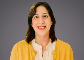 Portrait of a person with shoulder-length dark hair, smiling, wearing a white blouse and yellow cardigan, against a grey background.