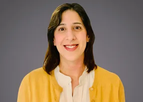 Portrait of a person with shoulder-length dark hair, smiling, wearing a white blouse and yellow cardigan, against a grey background.