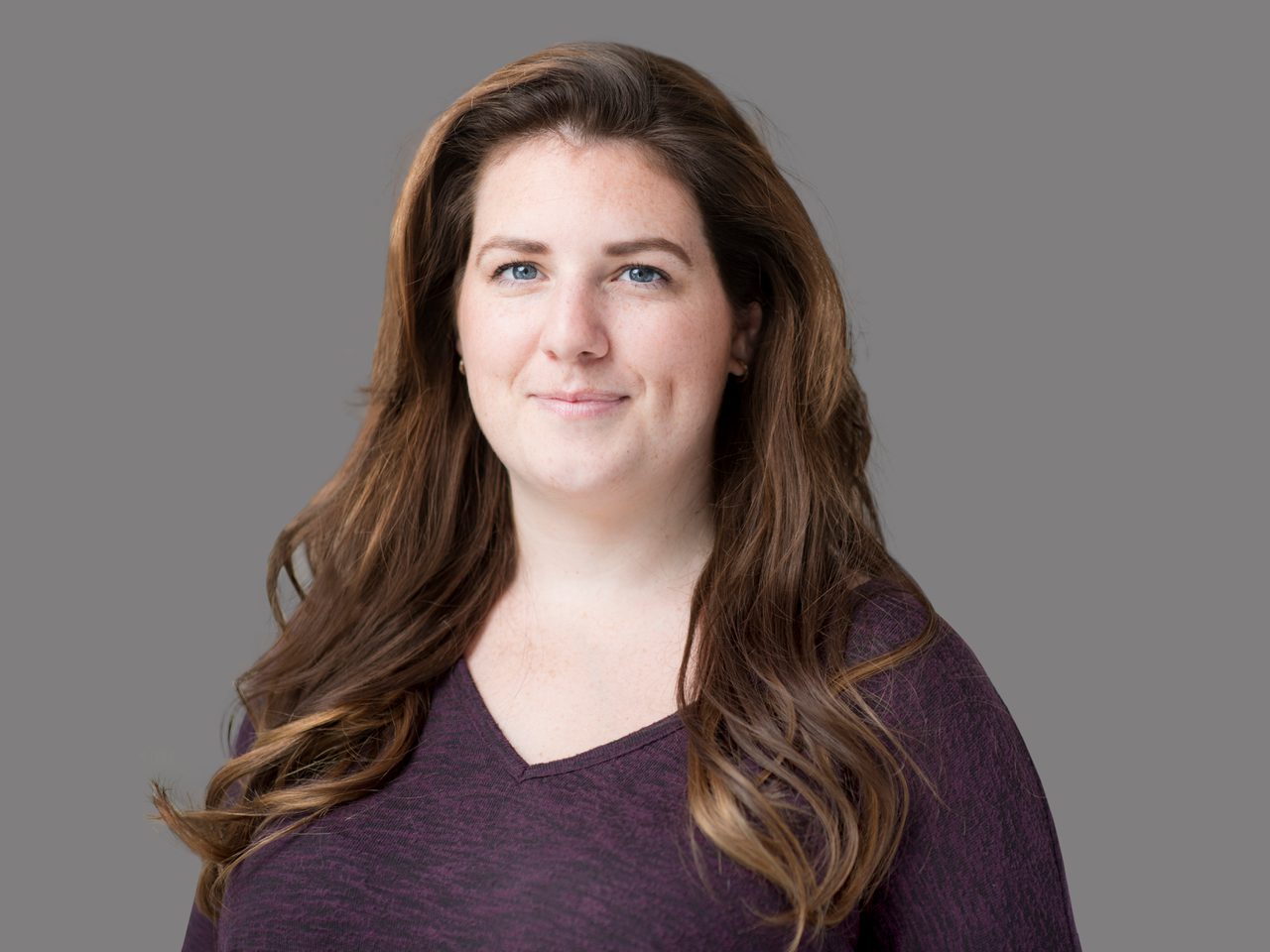Portrait of a person with long brown hair and blue eyes, wearing a maroon sweater, smiling softly at the camera against a gray studio background.
