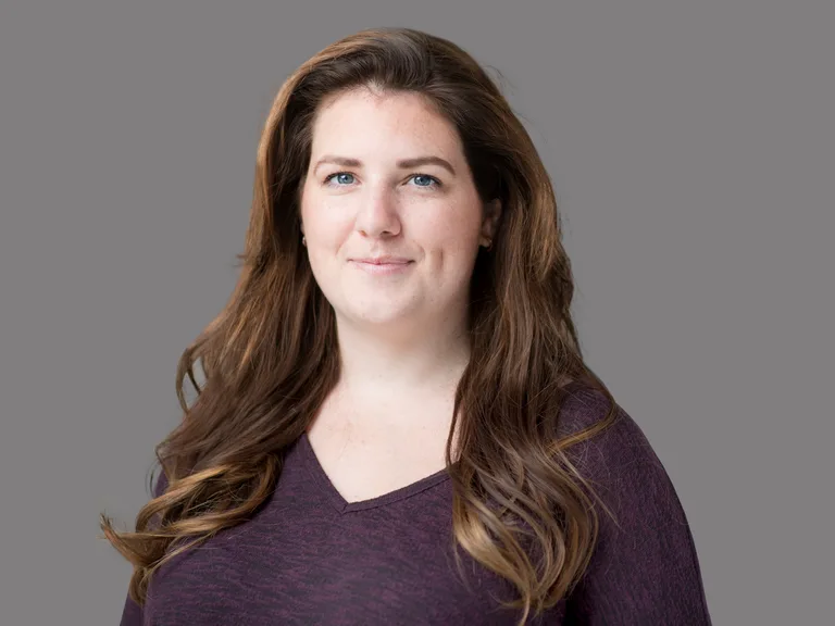 Portrait of a person with long brown hair and blue eyes, wearing a maroon sweater, smiling softly at the camera against a gray studio background.