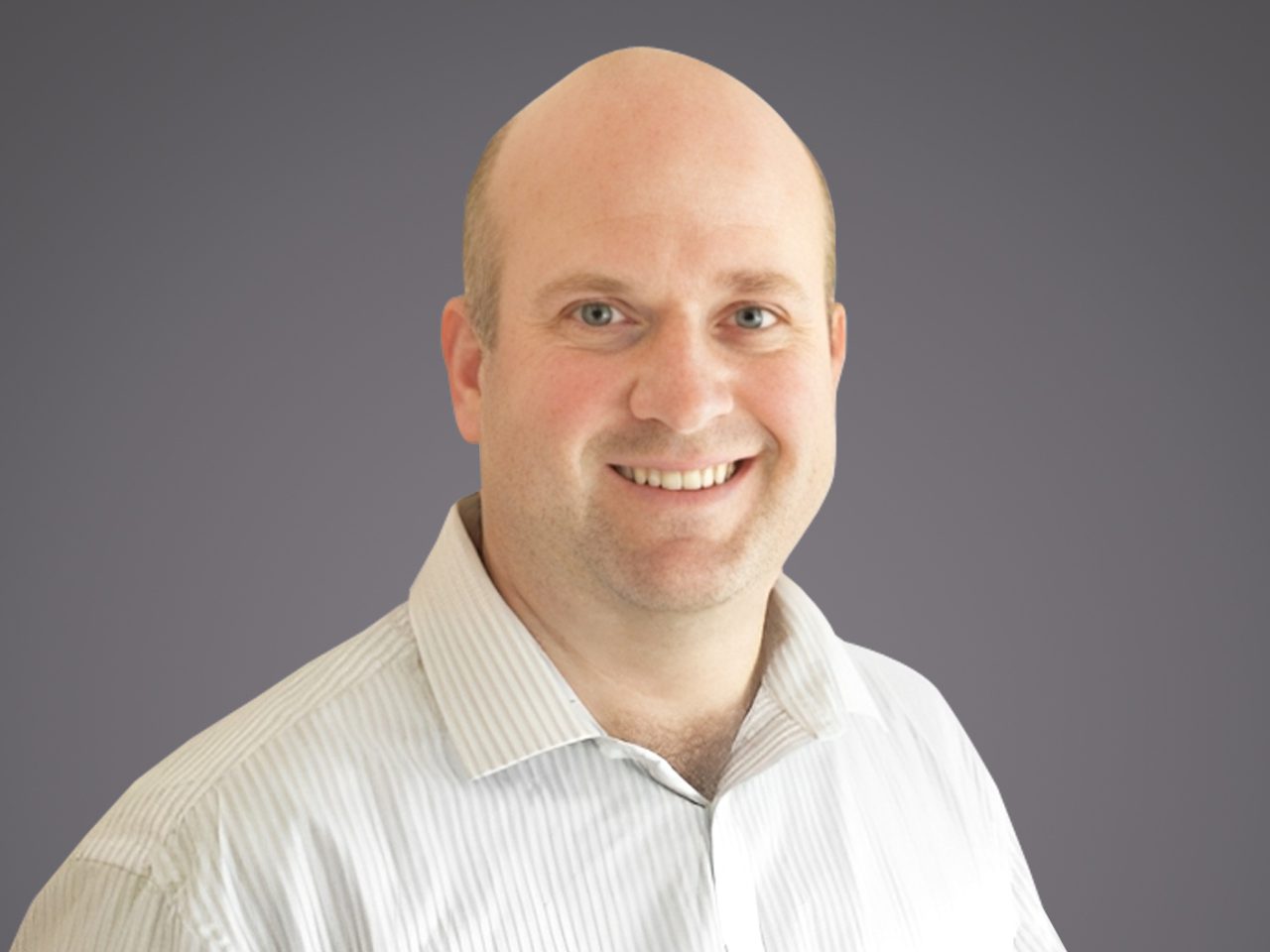 Head-and-shoulders portrait of a smiling, bald person in a light striped shirt against a neutral grey gradient background.