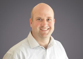 Head-and-shoulders portrait of a smiling, bald person in a light striped shirt against a neutral grey gradient background.
