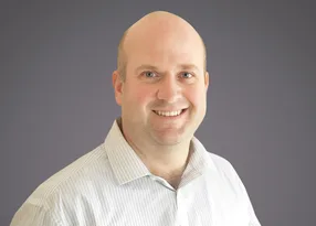 Head-and-shoulders portrait of a smiling, bald person in a light striped shirt against a neutral grey gradient background.