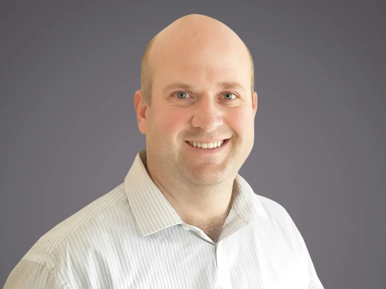 Head-and-shoulders portrait of a smiling, bald person in a light striped shirt against a neutral grey gradient background.