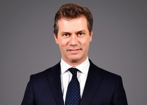 Personne en costume sombre, chemise blanche et cravate bleue à pois blancs, sourire neutre, fond gris.