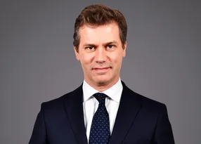 Professional head-and-shoulders portrait of a person in a dark suit, white shirt and navy dotted tie, against a neutral grey background.