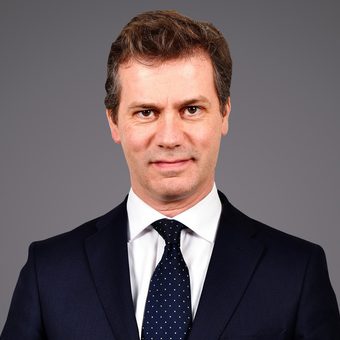 Personne en costume sombre, chemise blanche et cravate bleue à pois blancs, sourire neutre, fond gris.