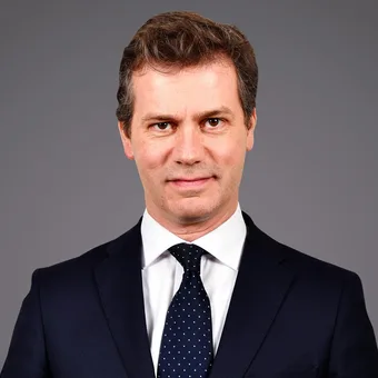 Personne en costume sombre, chemise blanche et cravate bleue à pois blancs, sourire neutre, fond gris.