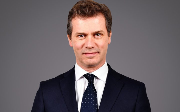 Personne en costume sombre, chemise blanche et cravate bleue à pois blancs, sourire neutre, fond gris.