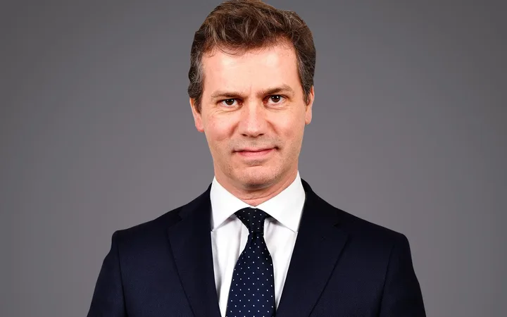 Personne en costume sombre, chemise blanche et cravate bleue à pois blancs, sourire neutre, fond gris.