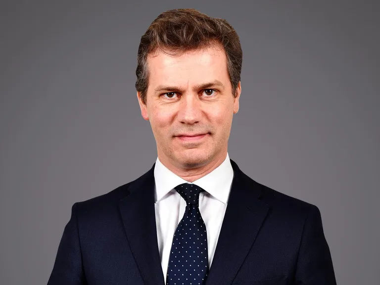 Person in dunklem Anzug, weißem Hemd und dunkelblauen Krawatte mit weißen Punkten, vor grauem Hintergrund, blickt ernst in die Kamera.