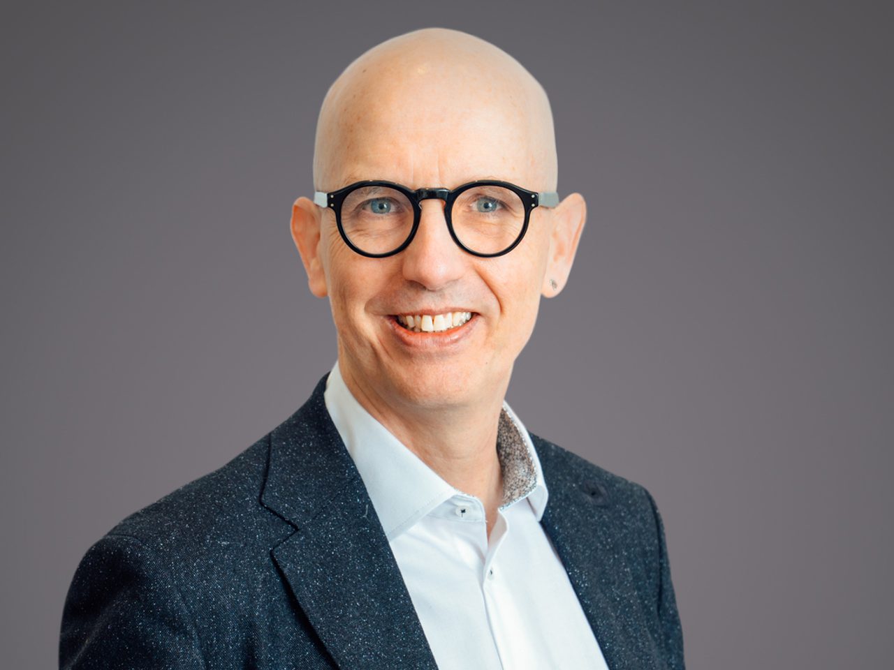 Portrait of a bald person with round black glasses, wearing a dark speckled blazer and light blue shirt, smiling against a neutral grey backdrop.
