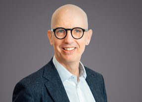 Portrait of a bald person with round black glasses, wearing a dark speckled blazer and light blue shirt, smiling against a neutral grey backdrop.