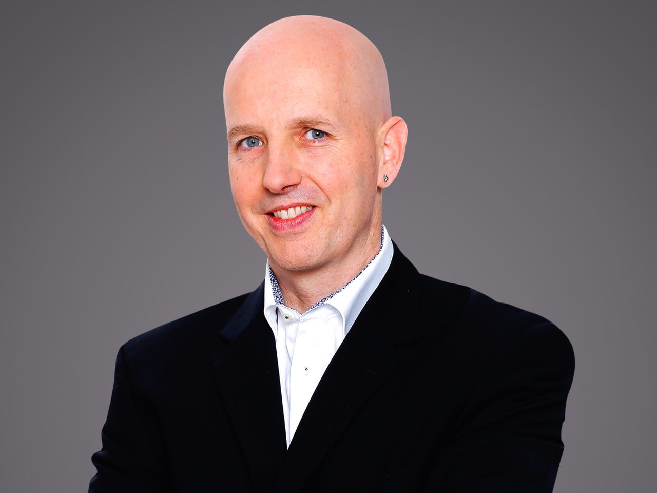 Bald person wearing a dark blazer and white shirt, smiling, with an earring, against a neutral grey studio backdrop.