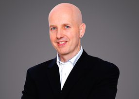 Bald person wearing a dark blazer and white shirt, smiling, with an earring, against a neutral grey studio backdrop.