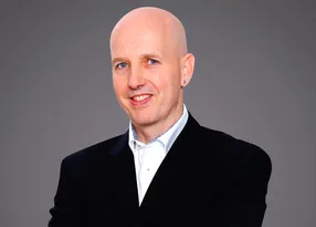 Bald person wearing a dark blazer and white shirt, smiling, with an earring, against a neutral grey studio backdrop.