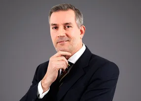 Portrait of a person in a dark suit and striped tie, with one hand on the chin, looking thoughtful at the camera against a plain grey backdrop.