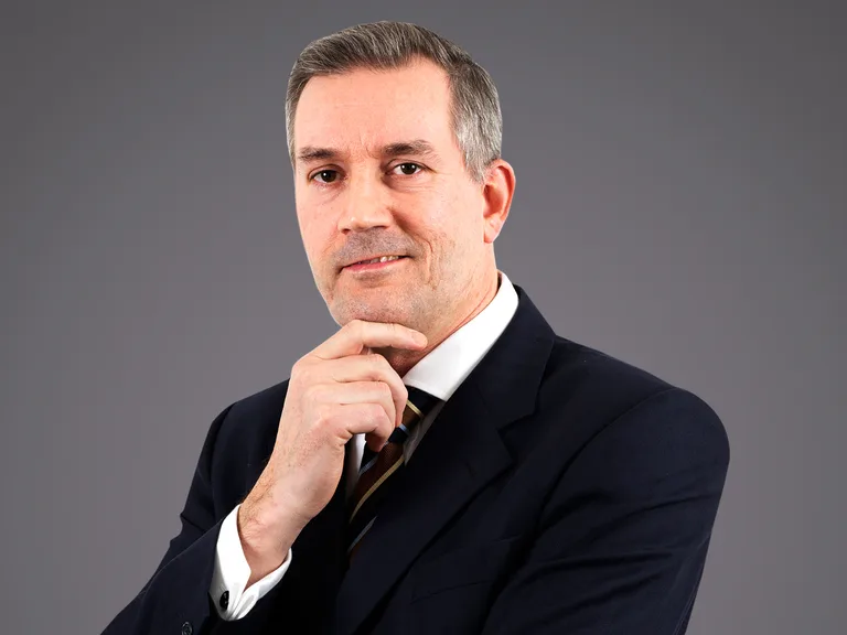 Portrait of a person in a dark suit and striped tie, with one hand on the chin, looking thoughtful at the camera against a plain grey backdrop.