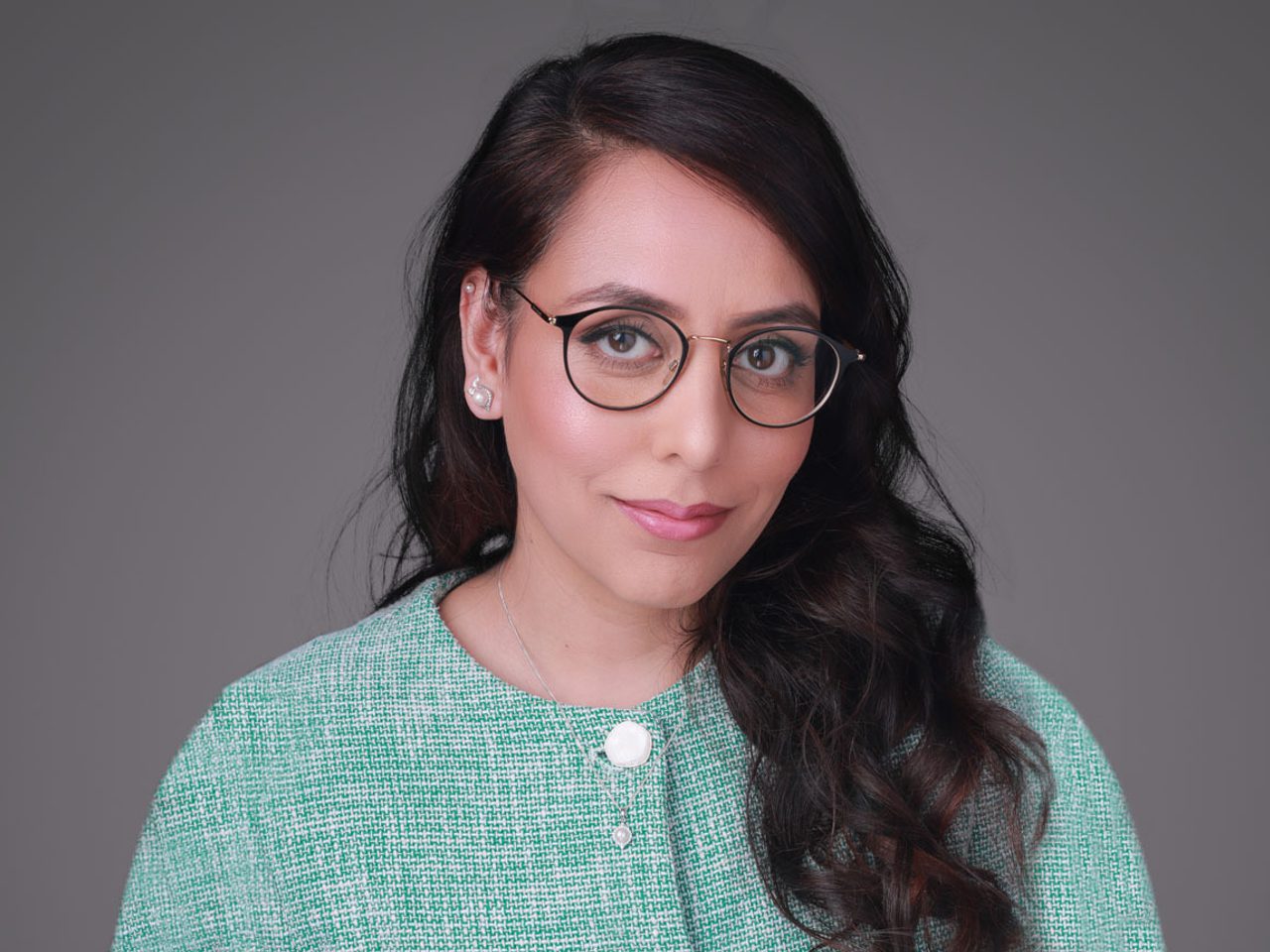Portrait of a person with long dark hair, round glasses, and a green textured top, smiling softly against a grey studio backdrop.