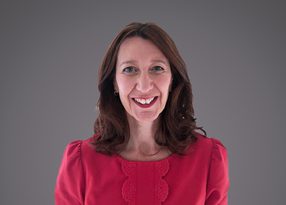 Smiling person with shoulder-length brown hair, wearing a pink blouse with lace trim, against a grey gradient background.