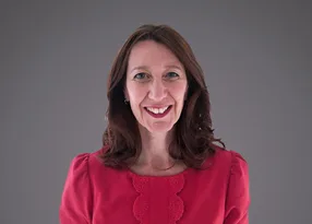Smiling person with shoulder-length brown hair, wearing a pink blouse with lace trim, against a grey gradient background.