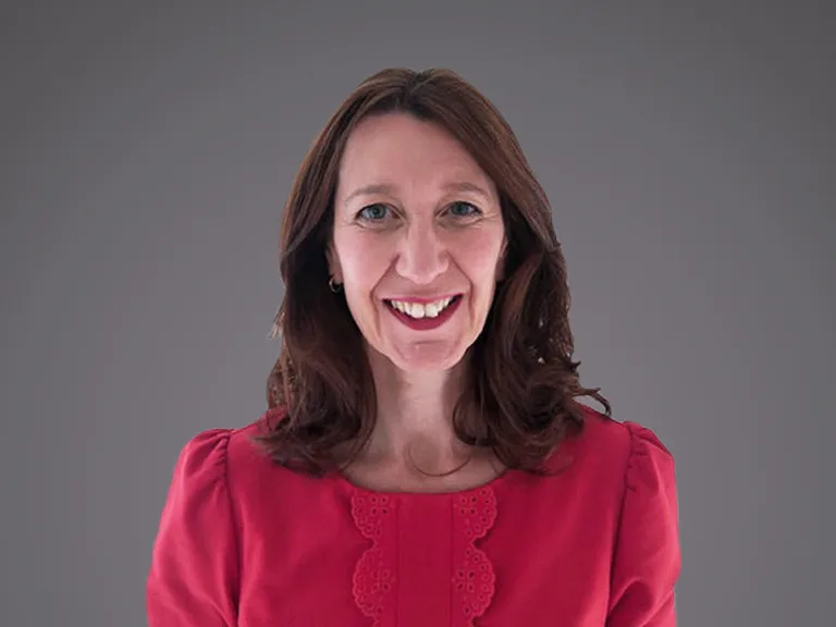 Smiling person with shoulder-length brown hair, wearing a pink blouse with lace trim, against a grey gradient background.