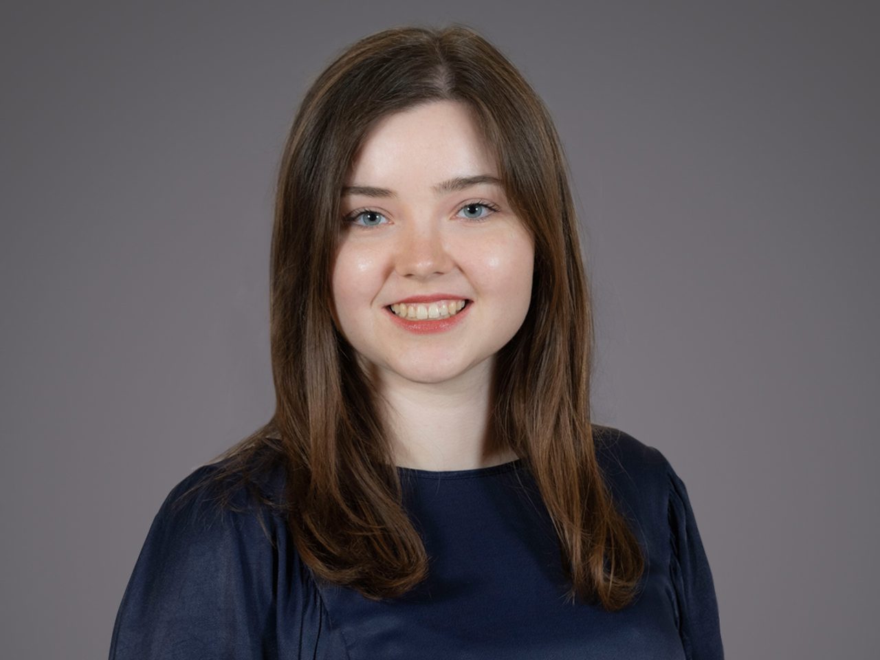 Portrait of a person with shoulder-length brown hair and blue eyes, wearing a navy top, smiling against a neutral grey backdrop.