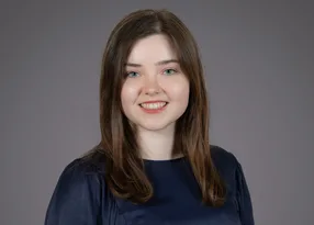 Portrait of a person with shoulder-length brown hair and blue eyes, wearing a navy top, smiling against a neutral grey backdrop.