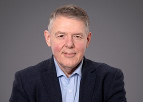 Portrait of a middle-aged person with short grey hair, wearing a light blue shirt and dark blazer, facing the camera against a grey studio backdrop.